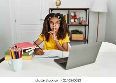 Young african american girl doing homework at home asking to be quiet with finger on lips. silence and secret concept. 