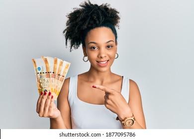 Young african american girl holding philippine peso banknotes smiling happy pointing with hand and finger 