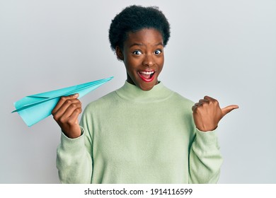 Young African American Girl Holding Paper Airplane Pointing Thumb Up To The Side Smiling Happy With Open Mouth 