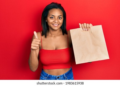 Young African American Girl Holding Take Away Paper Bag Smiling Happy And Positive, Thumb Up Doing Excellent And Approval Sign 