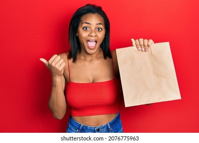 Young African American Girl Holding Take Away Paper Bag Pointing Thumb Up To The Side Smiling Happy With Open Mouth 