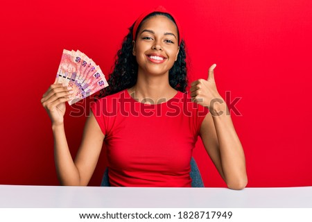 Young african american girl holding south african 50 rand banknotes sitting on the table smiling happy and positive, thumb up doing excellent and approval sign 