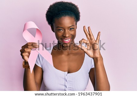 Young african american girl holding pink cancer ribbon doing ok sign with fingers, smiling friendly gesturing excellent symbol 