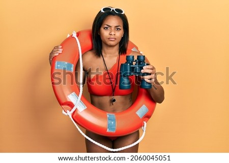 Young african american girl holding lifeguard floater using binoculars clueless and confused expression. doubt concept. 