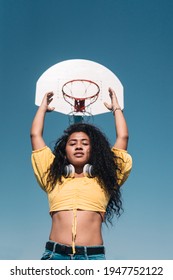 Young African American Girl Raising Her Arms Towards A Basketball Basket Looking At The Camera.