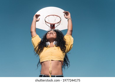 Young African American Girl Raising Her Arms Towards A Basketball Basket With Her Eyes Closed.