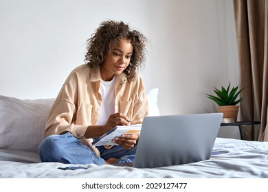 Young African American girl student studying virtual course, learning distance class taking web training, looking at laptop computer watching online webinar and writing notes sitting on bed at home.