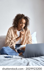 Young African American girl student studying virtual course, learning distance class taking web training, looking at laptop computer watching online webinar lecture sitting on bed at home.
