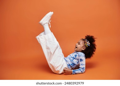 A young African American girl in a trendy outfit and hair clips poses on an orange background. - Powered by Shutterstock