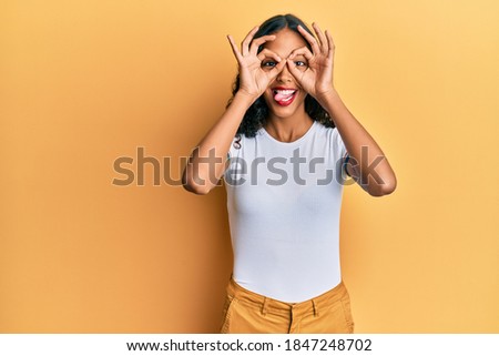 Young african american girl wearing casual clothes doing ok gesture like binoculars sticking tongue out, eyes looking through fingers. crazy expression. 