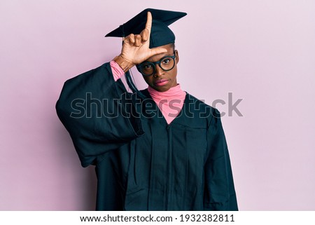Young african american girl wearing graduation cap and ceremony robe making fun of people with fingers on forehead doing loser gesture mocking and insulting. 