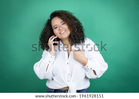 Young african american girl wearing white shirt over green background having conversation talking on the smartphone smiling happy pointing with hand and finger