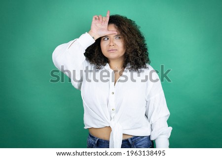 Young african american girl wearing white shirt over green background making fun of people with fingers on forehead doing loser gesture mocking and insulting.