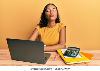 Young african american girl working at the office with laptop and calculator looking at the camera blowing a kiss on air being lovely and sexy. love expression. 