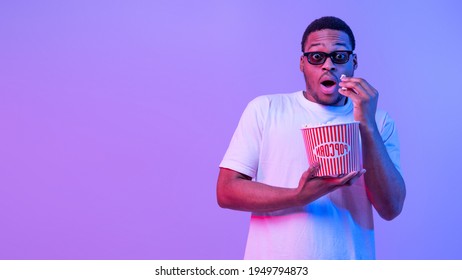 Young African American Guy In 3D Glasses Eating Popcorn While Watching Scary Movie In Cinema, Shocked Black Man Opening Mouth In Amazement, Standing In Neon Light On Purple Background, Panorama - Powered by Shutterstock