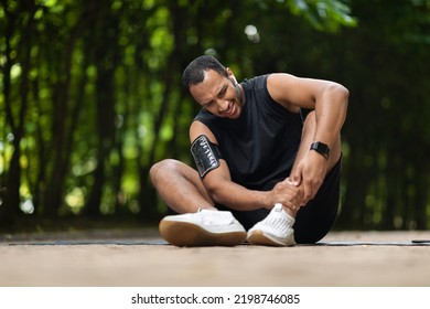 Young African American Guy Athlete In Pain Sitting On Fitness Mat In Public Park, Touching His Sore Ankle, Black Man Got Injured While Training Outdoors, Panorama With Copy Space