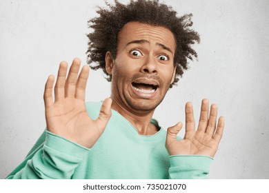 Young African American guy being in panic, looks terrified and troublesome, makes frightened gesture with palms, tries to defend himself from someone, asks stop it immediately. Emotional hipster