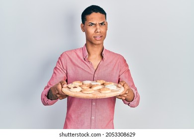 Young African American Guy Holding Tray With Cake Sweets Clueless And Confused Expression. Doubt Concept. 