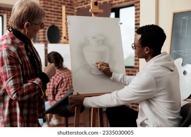 Young African American guy instructor teaching senior woman to draw during art class, teacher sitting at easel showing mature student how to hold and use pencil. Taking up drawing lesson in retirement - Powered by Shutterstock