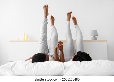 Young african american guy and lady in love lie on white bed in domestic clothes hold hands, raise legs up and relax in bedroom. Rest at free time and weekend, love, romance at home during covid-19