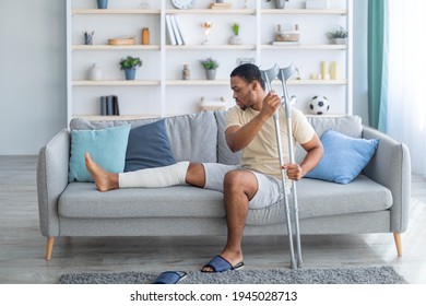 Young African American Guy With Plastered Leg Sitting On Sofa, Leaning On Crutches At Home. Millennial Black Man Suffering From Pain In Broken Limb, Resting On Couch, Full Length Portrait