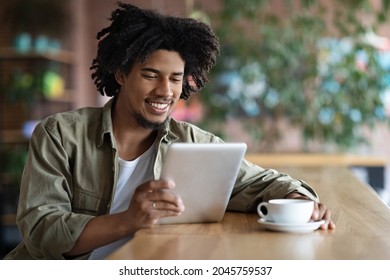 Young African American Guy Relaxing With Digital Tablet And Coffee In Cafe, Smiling Black Guy Browsing Social Media Or Reading News On Modern Gadget While Resting In Cafeteria, Free Space