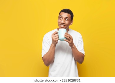 young african american guy in white t-shirt holds cup of coffee on yellow isolated background, man with drink looks at copy space
