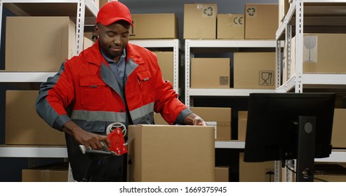 Young African American Handsome Post Male Worker In Uniform Packing Carton Box At Delievery Room In Post Office. Postman Closing Parcel With Sticky Tape. Mail Shipping Concept.