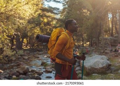 A Young African American Hiker Is Crossing A Small River Stream With His Back Pack And Walking Poles In The Mountains