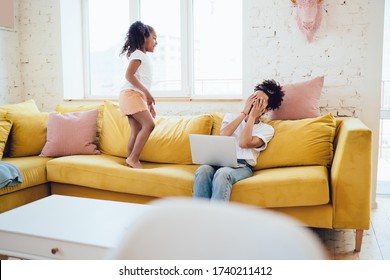 Young African American irritated woman with laptop covering face with hands in exhaustion while joyful daughter jumping on yellow sofa and looking at mother at home