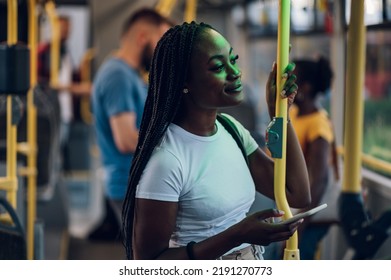 Young african american joyful woman standing on the bus during a night and using the smartphone and smiling while being illuminated by the city lights. Public transportation and people concept.