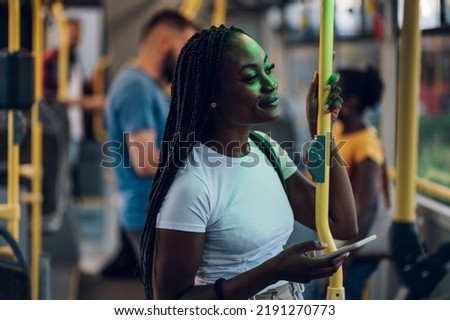 Young african american joyful woman standing on the bus during a night and using the smartphone and smiling while being illuminated by the city lights. Public transportation and people concept.