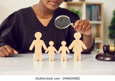 Young African American Judge In Black Gown With Magnifying Glass Looking On Family Wood Figure. Cropped Shot. Disputes Mediation, Children Custody By Parents After Divorce Court Decision