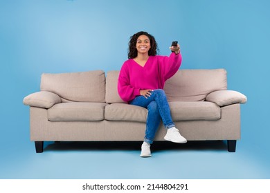 Young African American Lady With Remote Control Sitting On Couch And Watching TV On Blue Studio Background, Copy Space. Millennial Woman With Television Controller Switching Channels