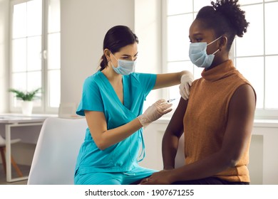 Young African American lady sitting at doctor's office and getting modern Covid 19 shot. Nurse in medical face mask and gloves holding syringe and giving female patient flu vaccine injection in arm