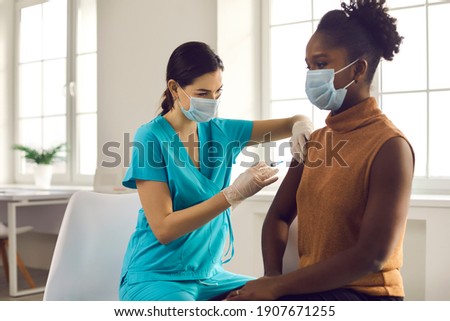 Young African American lady sitting at doctor's office and getting modern Covid 19 shot. Nurse in medical face mask and gloves holding syringe and giving female patient flu vaccine injection in arm