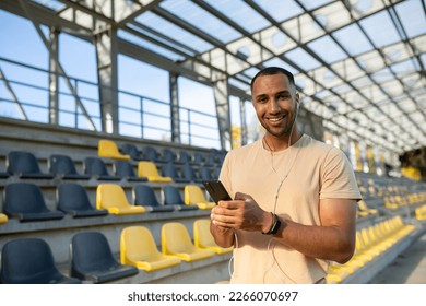 Young African American male athlete, coach, runner wearing headphones stands in stadium between bleachers, uses mobile phone. He looks at the camera, smiles. - Powered by Shutterstock