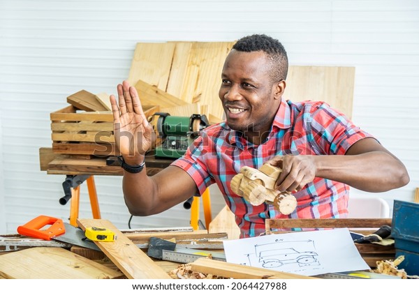Young african\
american male carpenter sitting at table showing model car wooden\
and drawing paper through tablet while video online chatting with\
customer or teaching online in\
factory