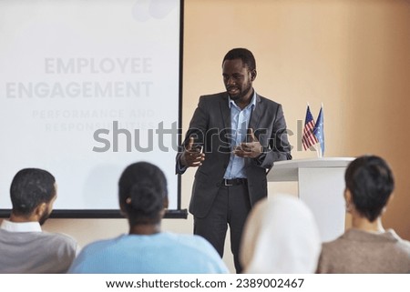 Young African American male deputy in formalwear speaking to audience while explaining them one of presentation points at conference