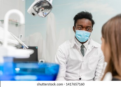 Young African American Male Doctor Dentist In A Medical Mask. Beautiful Girl Patient At Chair At Dental Clinic. Medicine, Health, Stomatology Concept. Dentist Conducts Inspection And Concludes