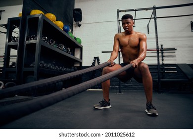 Young African American Male Exercising Inside A Gym. Topless Male Personal Trainer Using Rope To Strengthen His Arms. High Quality Photo