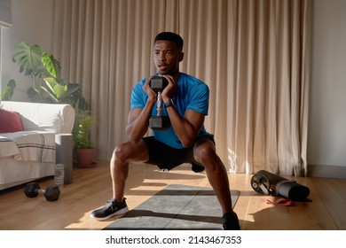 Young African American male exercising from home concentrating while squatting with a dumbbell in living room, workout at home - Powered by Shutterstock