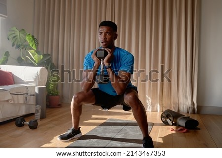 Young African American male exercising from home concentrating while squatting with a dumbbell in living room, workout at home