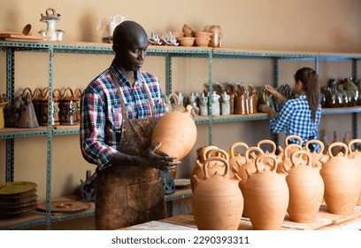 Young African American male hobbyist engaged in stoneware production in pottery workshop, looking at products - Powered by Shutterstock