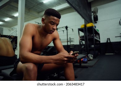 Young African American Male Looking At His Phone To Reply To An Online Message. Male Personal Trainer Looking At His Phone While Sitting Down In The Gym. High Quality Photo 