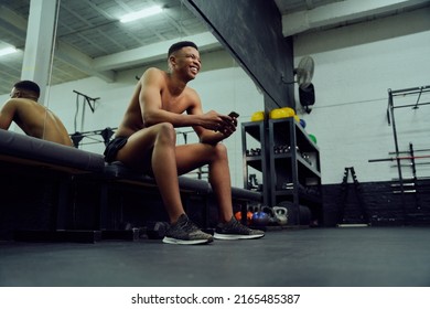 Young African American Male Looking At His Phone To Reply To An Online Message. Happy, Mixed Race Male Smiling At His Phone While Sitting Down In The Gym. High Quality Photo 