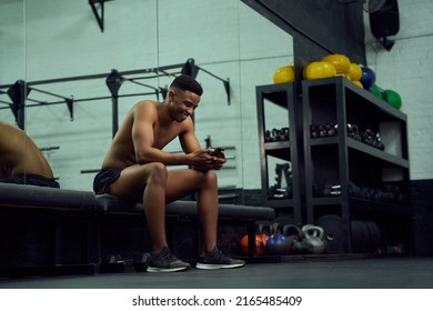 Young African American Male Looking At His Phone To Reply To An Online Message. Happy, Mixed Race Male Smiling At His Phone While Sitting Down In The Gym. High Quality Photo 