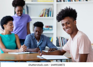 Young African American Male Student Learning At Desk At School With Teacher And Group Of Students At Classroom