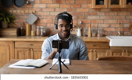 Young African American male vlogger influencer in headphones record video blog on smartphone at home. Millennial biracial man blogger talk speak shoot live broadcast on cellphone for social media.