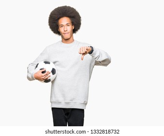 Young African American Man With Afro Hair Holding Soccer Football Ball With Angry Face, Negative Sign Showing Dislike With Thumbs Down, Rejection Concept